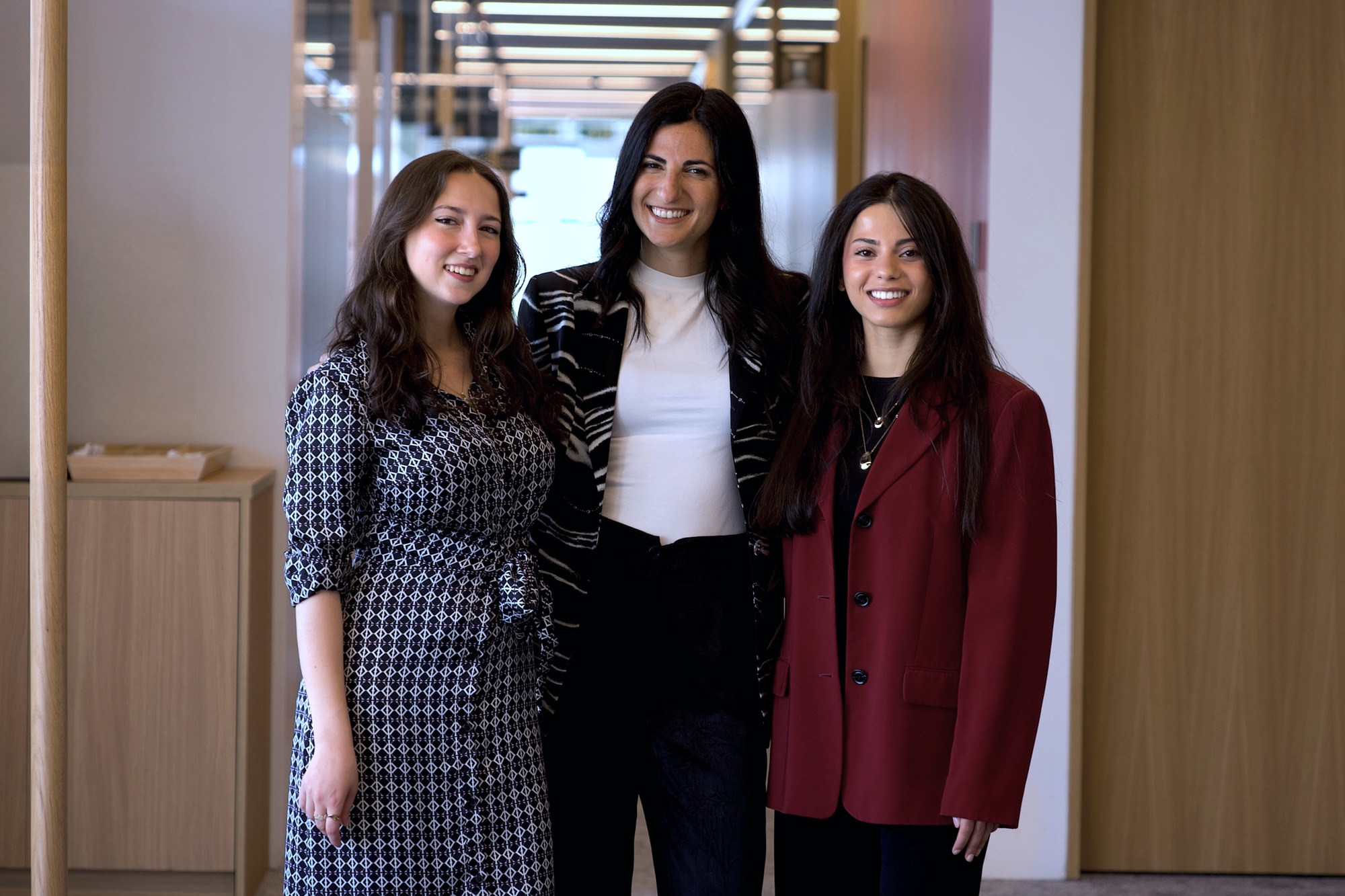 Three women standing together and smiling in a bright office corridor